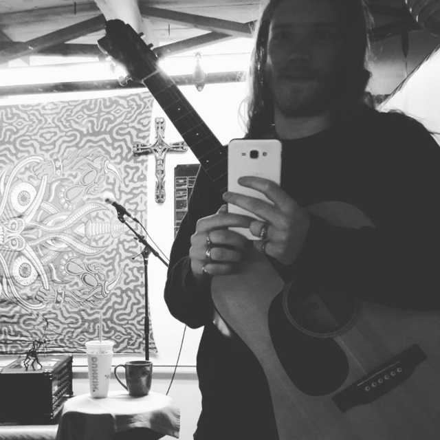 Walter, holding a guitar and a cell phone, taking a picture in the studio mirror,
		with a cross in the background, in black and white.